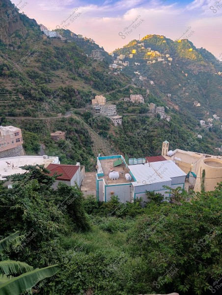 A scenic view of a mountainous village in a lush green area. The image displays houses scattered across hills covered with dense trees and vegetation. Winding roads run between the homes on the mountainside, with sunlit homes casting a golden glow near the peak. The sky is blue with thin white clouds.