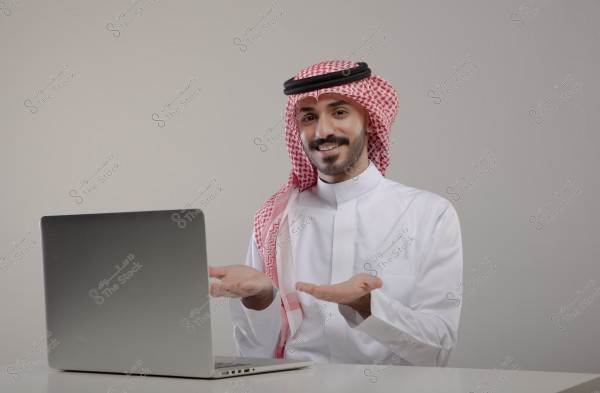An image of a man wearing traditional Gulf attire, including a white thobe, red checkered ghutra, and a black agal, sitting in front of a laptop on a white table. He is smiling and raising his hands in a relaxed expression. The background is simple and neutral-colored.