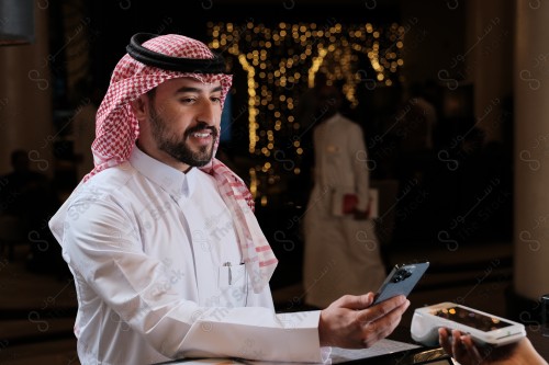 شاب سعودي في استقبال الفندق ، ويقوم بتعابير مختلفة باستخدام الجوال  ، ويقوم بدفع عبر جهاز الدفع الالكتروني