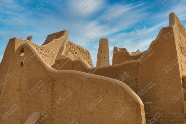 An image showing traditional mudbrick buildings with wavy clay walls and old architectural details under a clear blue sky. The architectural style resembles traditional Middle Eastern design, possibly from areas like Diriyah in Saudi Arabia.