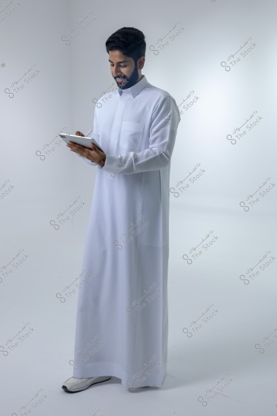 صورة لرجل يقف في استوديو ويرتدي ثوباً أبيض تقليدياً, قد يكون من السعودية. الرجل يحمل جهازًا لوحيًا وينظر إليه بابتسامة. الخلفية بيضاء بسيطة.