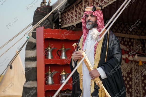 A portrait of a man wearing traditional Saudi attire, including a white thobe, a shiny black bisht with gold embroidery, a red and white shemagh, and a black agal. He stands in front of a traditional decorated tent with a red shelf beside him that holds a collection of Arabic silver coffee pots.