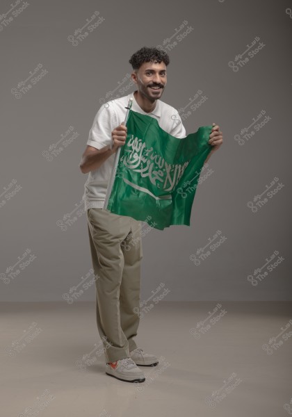 A young man holding the flag of his country, Saudi Arabia, standing with a smile, wearing a white shirt, beige pants, and sports shoes. The green flag with its emblem is clearly visible.