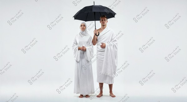 An image of a man and a woman standing side by side. The man is dressed in a white ihram garment and is holding an umbrella over them. The woman is wearing a long white robe and a hijab covering her hair. Both are barefoot, standing against a white background.