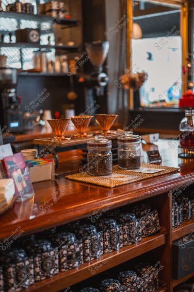 An indoor shot of a coffee shop featuring three copper coffee filters on a wooden counter, with two glass jars containing coffee beans and closed tins on a wooden shelf, with some utensils and decor visible in the background.