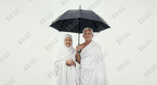 An elderly couple dressed in white ihram garments stands under a black umbrella. The woman is wearing a hijab with her ihram, while the man has wrapped the ihram around himself. They appear to be preparing for the Hajj pilgrimage in Saudi Arabia.