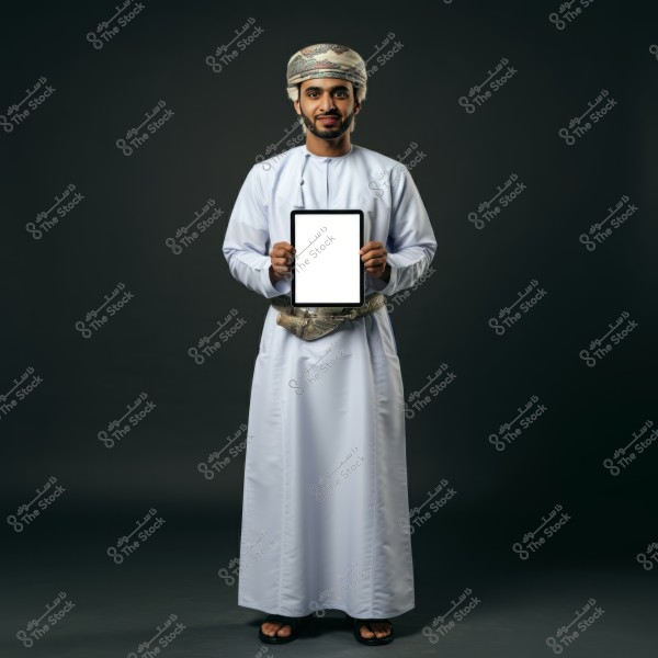 A portrait of a man standing against a dark background, wearing traditional Omani attire in white with a belt featuring a ceremonial dagger. The man wears an Omani turban and holds a tablet with a blank screen in front of him.