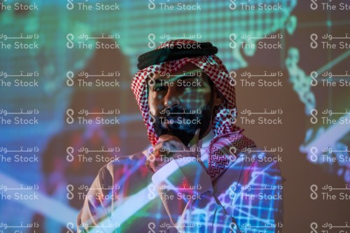 رجل سعودي يرتدي الزي السعودي التقليدي يضع يده على دقنه يفكر أثناء متابعة سوق الأسهم بينما تنعكس أضواء الشاشة عليه