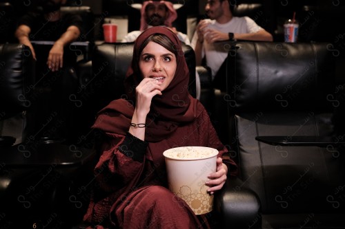 A Saudi girl in the cinema has interactions while watching the movie, she wears an abaya, and popcorn and a drink appear in front of her