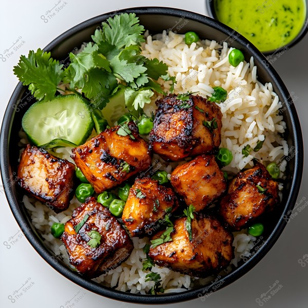 An image of a dish featuring browned, grilled tofu pieces on a bed of white rice. The dish is garnished with fresh green peas and cilantro leaves. A slice of cucumber is also visible, along with a small bowl of green sauce placed beside the dish.