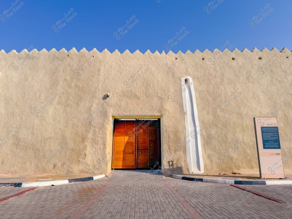 بوابة خشبية كبيرة ضمن جدار قلعة قديمة, مع لافتة توضح معلومات تاريخية, والسماء زرقاء صافية في الخلفية.