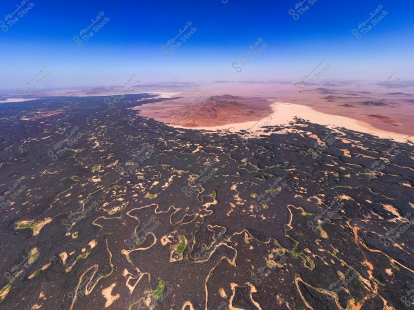 Aerial view of an expansive desert landscape featuring black volcanic hills interspersed with sandy patches and rugged rock formations. A range of mountains is visible on the horizon under a clear blue sky. The scene appears natural and vast, reflecting the geographical diversity of the desert.