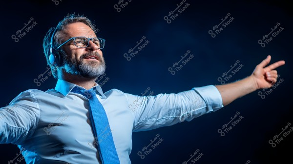 صورة لرجل يرتدي سماعات رأس ونظارات, يرفع يديه مشيراً بإحدى يديه, يرتدي قميصًا أزرق مفتوح الياقة وربطة عنق زرقاء. الخلفية زرقاء داكنة, والإضاءة موجهة على وجهه وملابسه مما يضيف إحساسًا دراميًا للمشهد.
