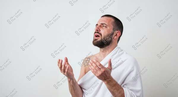 A portrait of a bearded man standing against a white background, appearing to be in a state of contemplation or prayer. He is draped in a white fabric, with a tattoo of a flower on his left shoulder. His hands are raised forward as if in invocation or supplication.