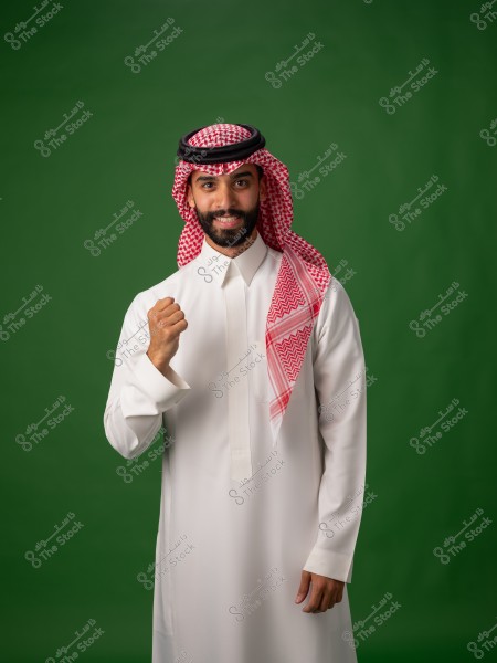 Portrait of a man standing in front of a green background. The man is wearing traditional white attire with a red and white checked headscarf and black agal, suggesting he may be from Saudi Arabia. He is expressing pride or enthusiasm with a clenched fist and is smiling.