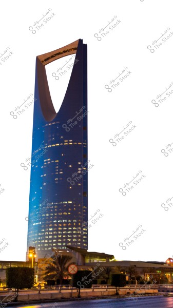 An image of the Kingdom Tower in Riyadh, Saudi Arabia. The tower is shown with its distinctive height and shiny blue color while the windows are lit on the upper part of the tower, along with the wide arch-like top. At the bottom, some trees, plants, and a road are visible in the foreground.