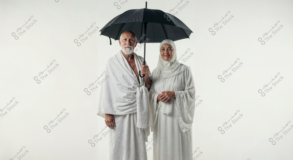 Image of a man and woman standing under a black umbrella. The man is wearing a white Ihram, which is a garment worn during the Hajj or Umrah pilgrimage. The woman is dressed in a traditional white dress with a headscarf. The image appears to represent a religious or cultural theme.