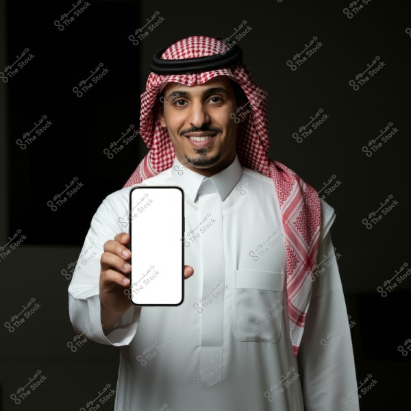 A portrait of a man wearing traditional Saudi attire, including a white thobe and red and white checkered shemagh, holding a smartphone with a blank white screen. The background is dark, highlighting the person and the phone. The image suggests showcasing or presenting something on the phone.