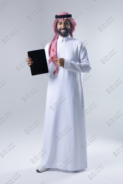 Image of a man wearing traditional Saudi attire consisting of a white thobe, red and white ghutra, and a black agal. The man is smiling and holding a tablet in his right hand while pointing at it with his left finger.