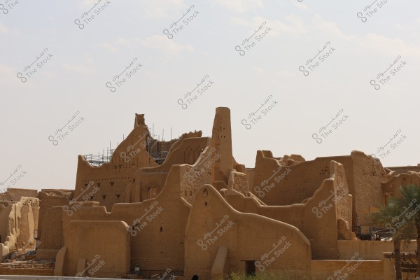 An image depicting ancient ruins made of mud located in a desert area. The structures are traditional mud buildings with intricate geometric shapes, and some scaffolding is visible on top, indicating restoration work. The sky is clear with a few scattered clouds, and a palm tree is visible in the lower right corner.