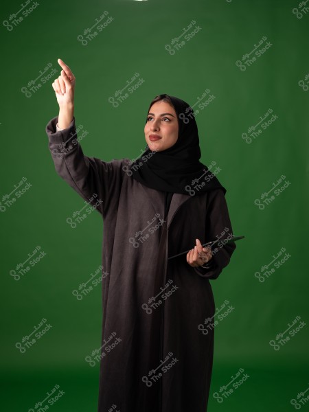 صورة شخصية لامرأة تقف أمام خلفية خضراء. ترتدي عباءة سوداء وحجاباً وتبدو وكأنها تشير بأصبعها إلى شيء في الهواء. تحمل في يدها جهازاً لوحياً.