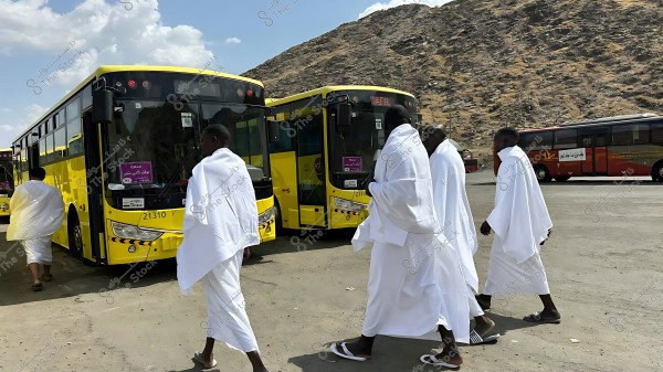 Pilgrims wearing white Ihram garments walking towards yellow buses designated for transport in Mecca, with mountains in the background.