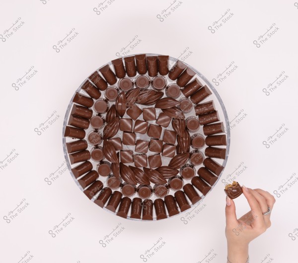 An image of a large circular platter containing a variety of chocolates arranged in concentric circles. A hand is holding one piece of chocolate from the platter, delicately at the lower right part of the image.