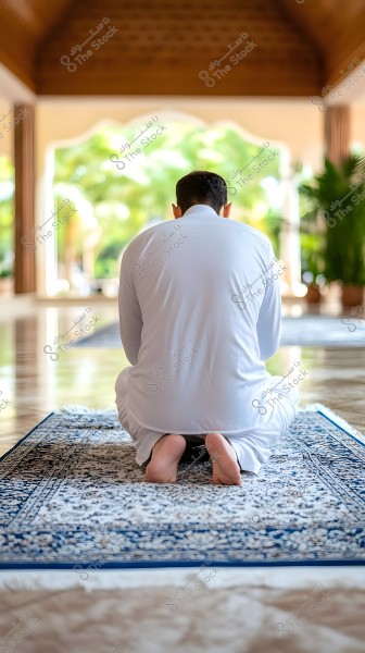 A person wearing a white thobe, sitting on a blue and white patterned prayer rug inside an open mosque. The scene is viewed from behind, with a natural backdrop outside the window featuring green trees and sunlight streaming in.