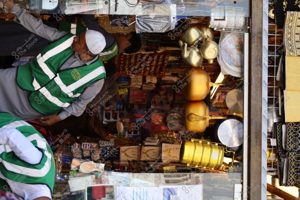 The image shows a shop displaying a variety of goods including metallic pots and golden-colored trays, as well as some fabrics. Two individuals stand in front of the shop wearing green vests with Arabic text and one wearing a white cap. The shelves are filled with various products such as jewelry made from gold or brass, and the background contains bags and household items.