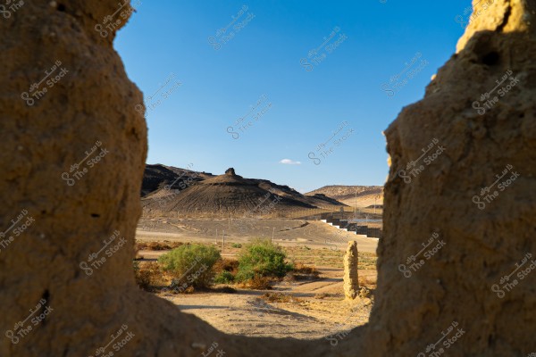 مشهد طبيعي يظهر من خلال فتحة في صخور طينية, يضم تلالًا بنية اللون مع بعض النباتات الخضراء المتفرقة في الأمام. السماء زرقاء صافية, وتظهر بعض الجدران والممرات الطينية في الخلفية.