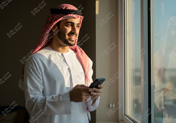 The image shows a person standing by a window, holding a mobile phone, wearing a traditional white thobe and a red and white ghutra with a black agal. The window reflects his image, and natural light illuminates his smiling face.