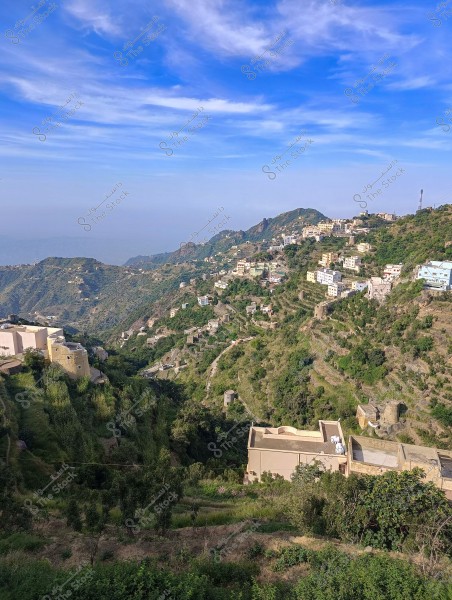 A scenic view of green hills with white houses and small buildings scattered across the slopes. Narrow roads wind between the houses, offering a stunning view of the valley. The sky is blue with light white clouds, creating a sense of tranquility and natural beauty.