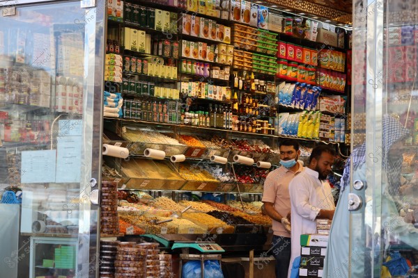 A small shop filled with various products such as spices, nuts, oils, herbs, and teas. The shelves are colorful with different packaging, and two men are seen behind the shop counter, one wearing a shirt and the other in traditional Arab attire. Another person in a headscarf and kandura is walking near the glass door of the shop.