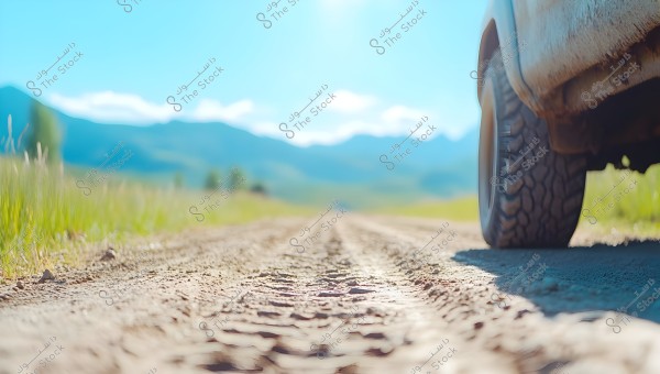 An image of a car driving on a dirt road in a rural area, with the car\'s tire prominently in the foreground. The road is bordered by green grass extending towards distant mountains under a clear blue sky with a few clouds.