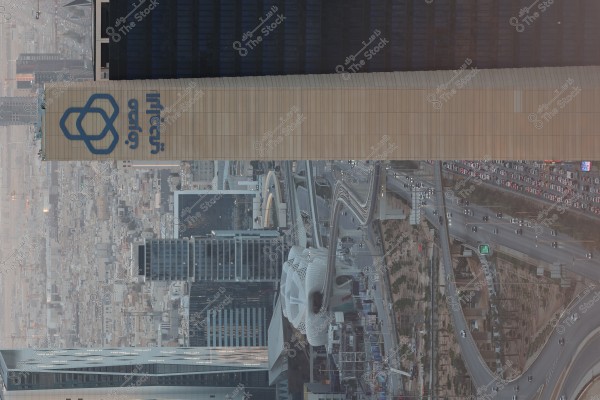 منظر جوي لأفق المدينة يظهر مبنى طويل مع شعار على جانبه الأيسر باللغة العربية. يمكن رؤية حركة المرور الكثيفة على الطرق السريعة التي تمر بين المباني التي تتنوع في ارتفاعاتها. تغطي المباني والأبراج زجاجية الجانب معظم المساحة في الصورة, وهناك مشاهد من العمارة الحديثة والطرق المتعرجة فيما يبدو أنها مدينة كبيرة في المملكة العربية السعودية.
