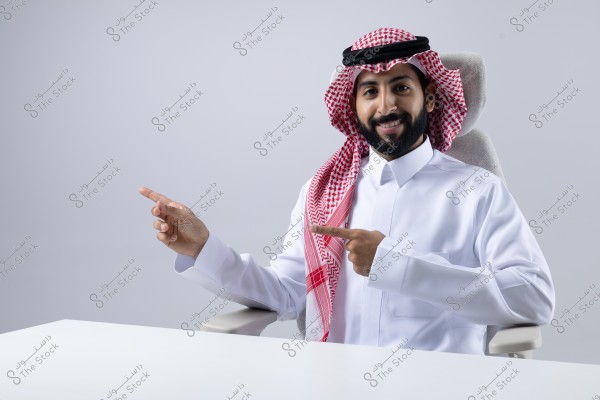 A portrait of a man sitting on a chair wearing a white thobe, red shemagh, and a black agal, pointing with his hands to the left against a grey background. He appears smiling and relaxed.