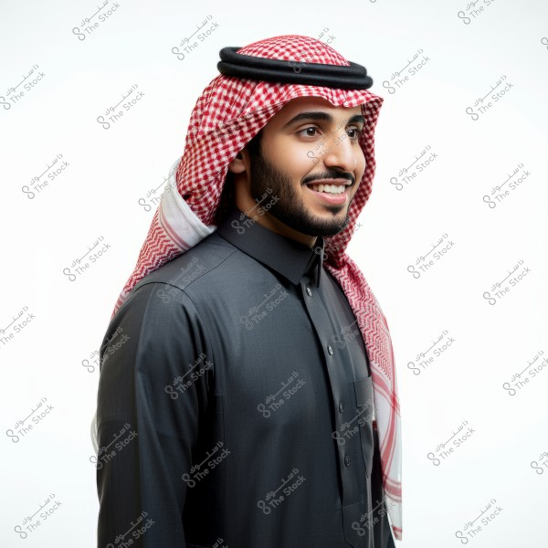 A portrait of a young man wearing traditional Saudi attire, consisting of a black thobe and a white and red checkered ghutra held in place with a black agal. The man is smiling confidently, set against a clean white background.