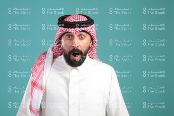 بورتريه رجل سعودية يرتدي الثوب ويقف أمام خلفية تركوازية ، مندهش ، تعابير وجه تدل على الاندهاش ، رجل متفاجئ