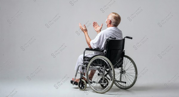 A man is seated in a wheelchair in a side view, wearing a white robe and a face mask. The man has his hands raised upwards as if in prayer or meditation. The background is white and neutral, drawing full attention to the person in the image.