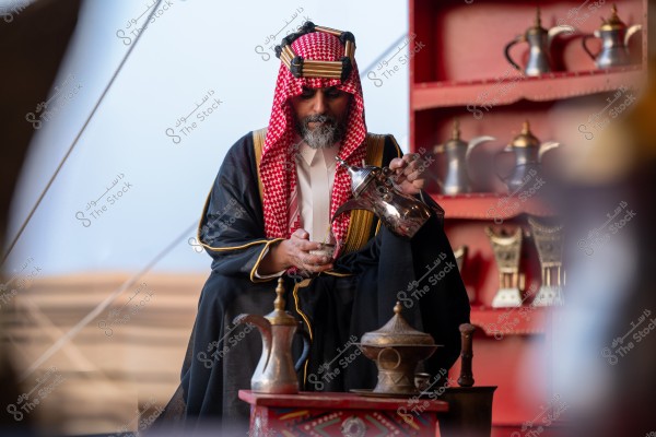 A man wearing traditional Arab clothing with a red checkered keffiyeh and agal, sits and pours Arabic coffee from a silver dallah into a small cup. In the background, there is a shelf with various dallahs and decorative cups. The setting appears to be a traditional gathering place in the desert with sand and an intricately decorated table in front of him.