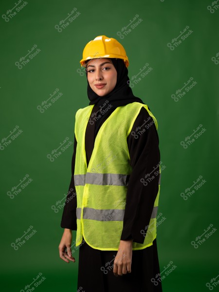 The image shows a woman wearing a yellow safety helmet and a high-visibility vest over a black abaya. She is standing in front of a green background, looking at the camera with a slight smile.