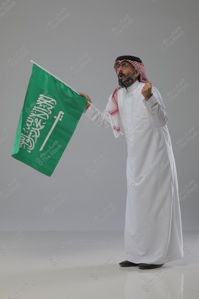 An image of a man wearing traditional Saudi attire, including a white thobe, a red and white keffiyeh, and a black agal. He is holding the Saudi Arabian flag in his right hand and appears to be in an enthusiastic posture, raising his left fist in a gesture of support or cheering.