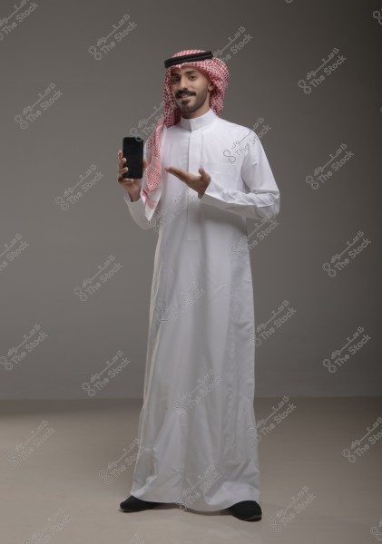 A portrait of a man wearing a white thobe and traditional headwear known as a ghutra and agal, smiling and pointing towards a smartphone he holds in his other hand. He appears to be from Saudi Arabia based on his traditional attire. The background is neutral and simple.