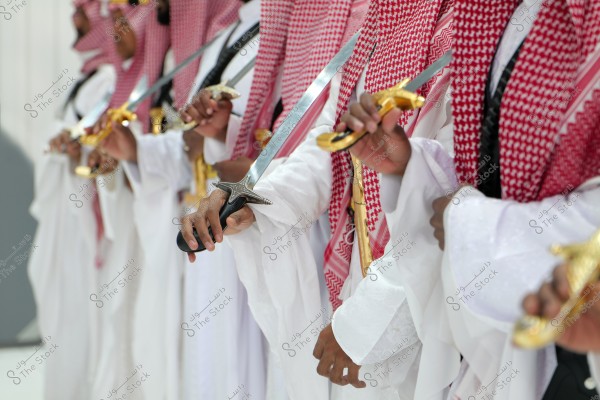 A group of men wearing white thobes and red-and-white ghutras are holding intricately decorated swords. The image represents an aspect of traditional cultural heritage, possibly related to Saudi Arabia based on the clothing style.