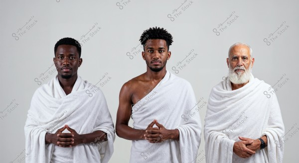 An image of three men standing side by side wearing white Ihram, the traditional clothing for men during the Hajj and Umrah pilgrimage in Islam. They are set against a simple gray background, with serious expressions and their hands clasped in front of them.