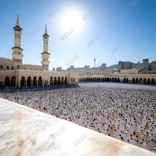 تظهر في الصورة ساحة المسجد الحرام في مكة, المملكة العربية السعودية, مكتظة بالناس يرتدون ملابس إحرام بيضاء بينما يقومون بأداء الصلاة. يظهر في الخلفية برج الساعة الشهير ومبانٍ شاهقة أخرى, وتظهر السماء زرقاء نقيّة والشمس تسطع في الأعلى.