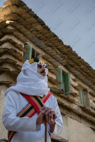 A snapshot of a Saudi man wearing traditional costume on the founding day, standing in front of a mud building and leaning on a stick, traditional costume, foundation day, ancient ancient buildings