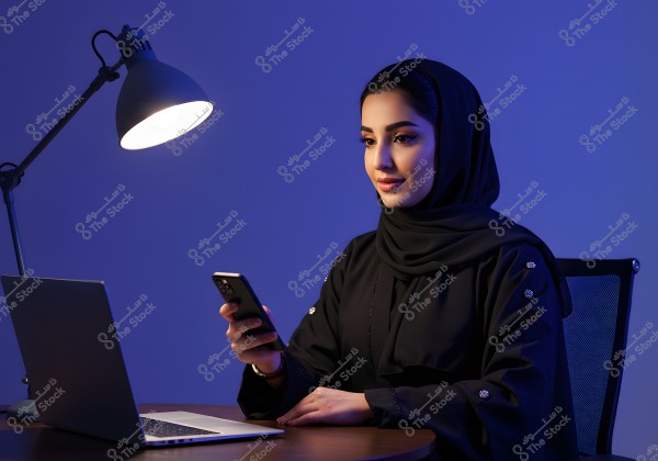 An image of a woman wearing a hijab and a black abaya working at a desk. In front of her is a laptop, and she is using a mobile phone. The lighting is dim, with a desk lamp illuminating the space. The background is dark blue, creating a calm and focused atmosphere. The attire suggests a style from the Gulf region.