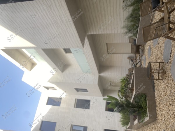 Image of a modern building facade in white with a clear blue sky in the background. At the bottom, there is an outdoor courtyard paved with pebbles, surrounded by several wooden chairs neatly arranged around a table. The courtyard is decorated with green plants placed in planters.