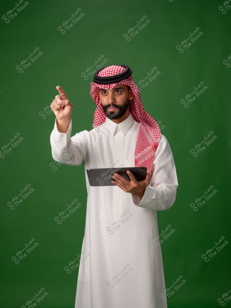 A portrait of a man wearing traditional Saudi attire, consisting of a white thobe, a red and white checkered keffiyeh, and a black agal, standing against a green background. He is holding a tablet in one hand and pointing upwards with his index finger in the other, appearing to be in a contemplative or instructive pose.